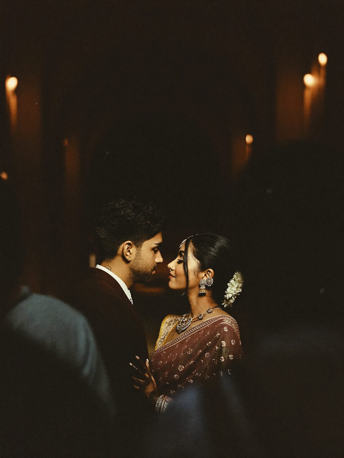 Couple nose to nose in a pocket of warm light, seen between blurred guests