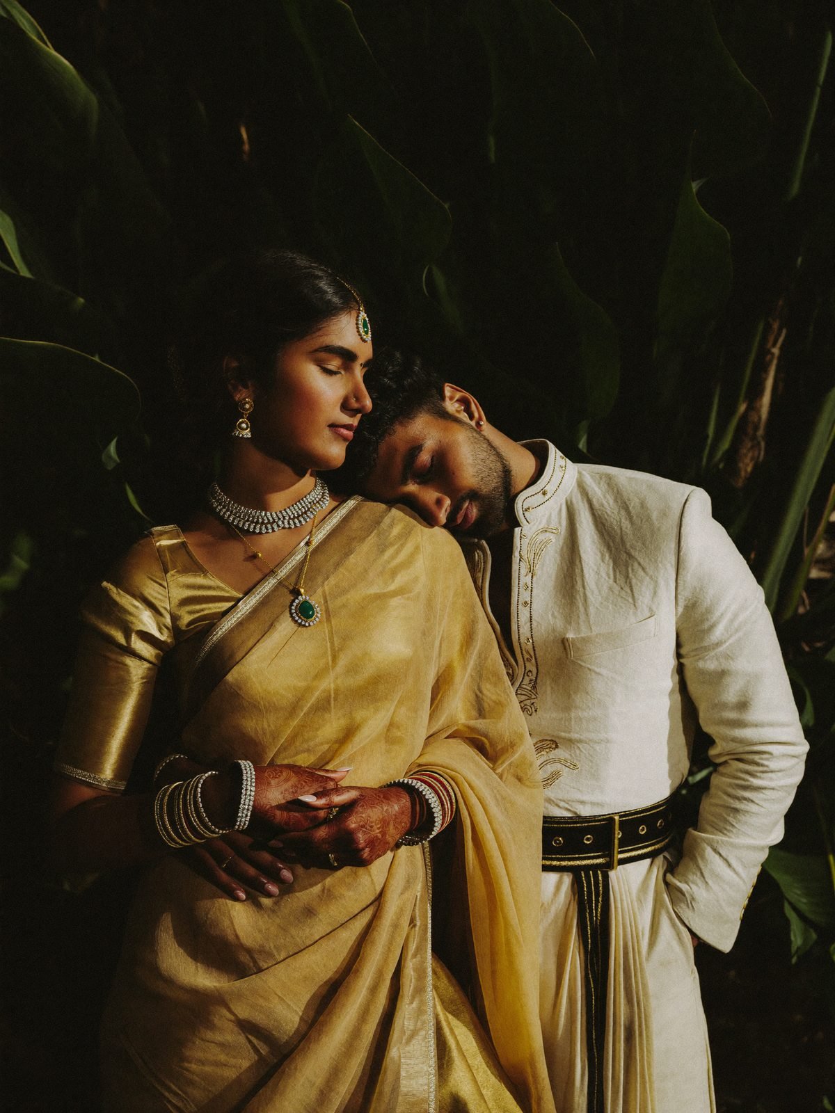 Kannadiga bride in gold Kanjeevaram with diamond jewellery embraced by the groom against dark foliage