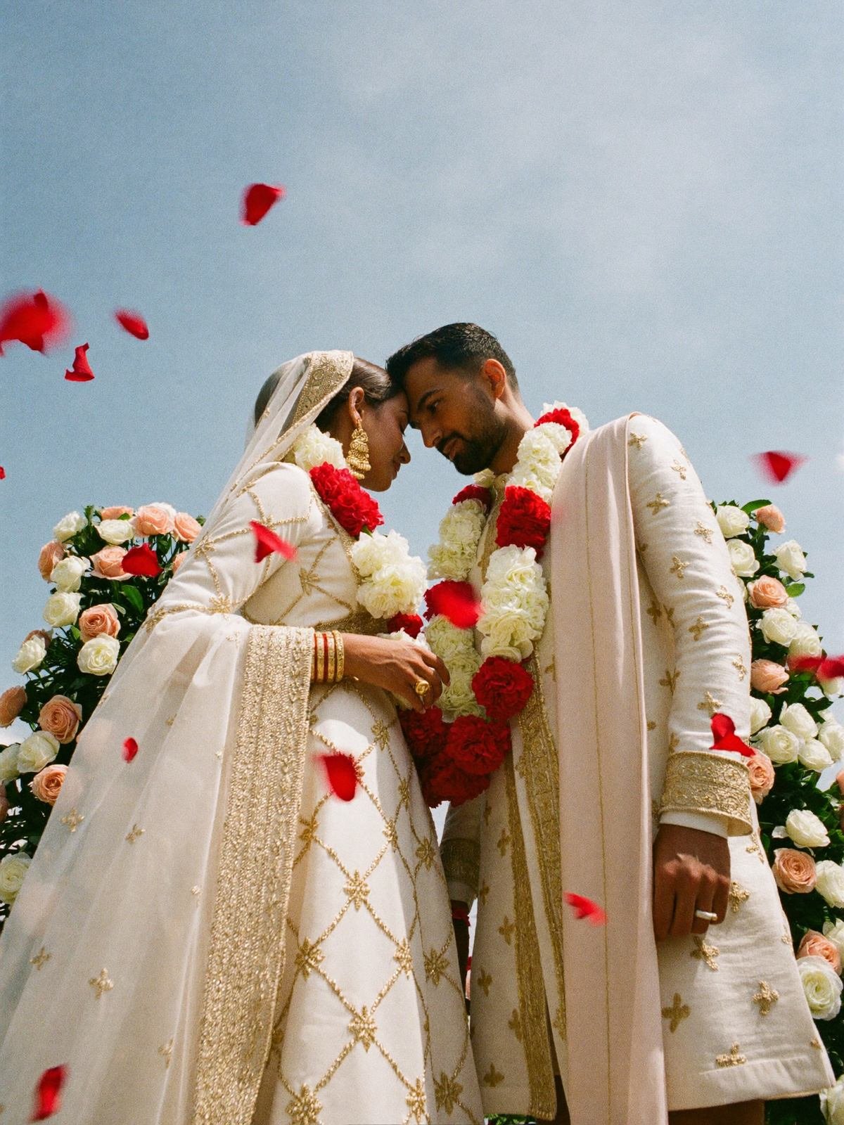 Couple in ivory couture with red garlands photographed low against an open sky with falling petals